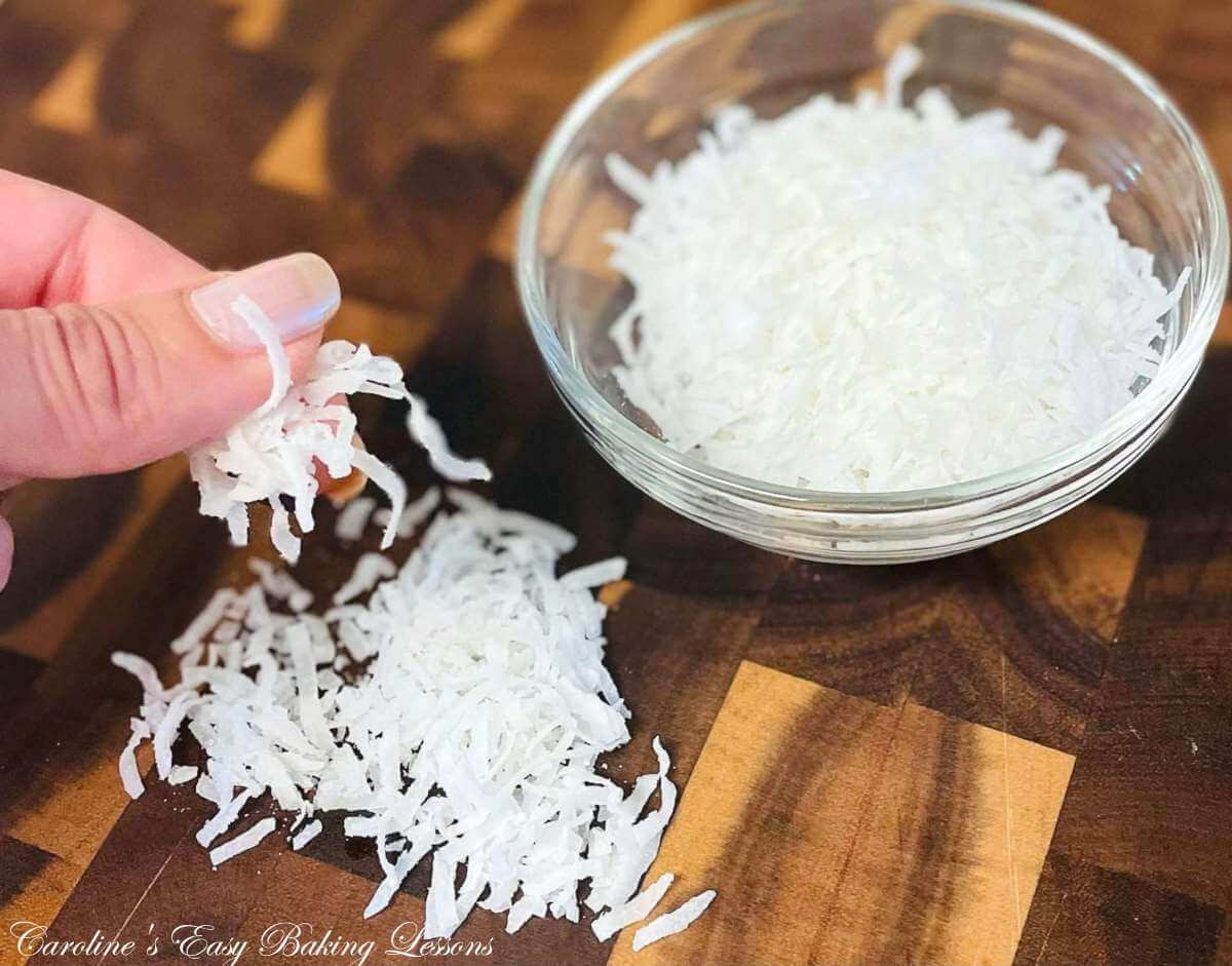 Female hand with long nails, holding US shredded coconut pieces between fingers an don dark wooden chopping board below.