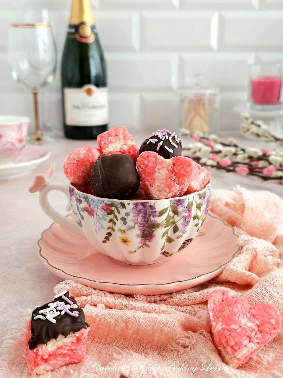 Close shot of a pink and floral teacup and saucer filled with pink and white coconut ice, heart shaped and chocolate coated pieces, with pink napkin, champaigne bottle and flowers to the background.