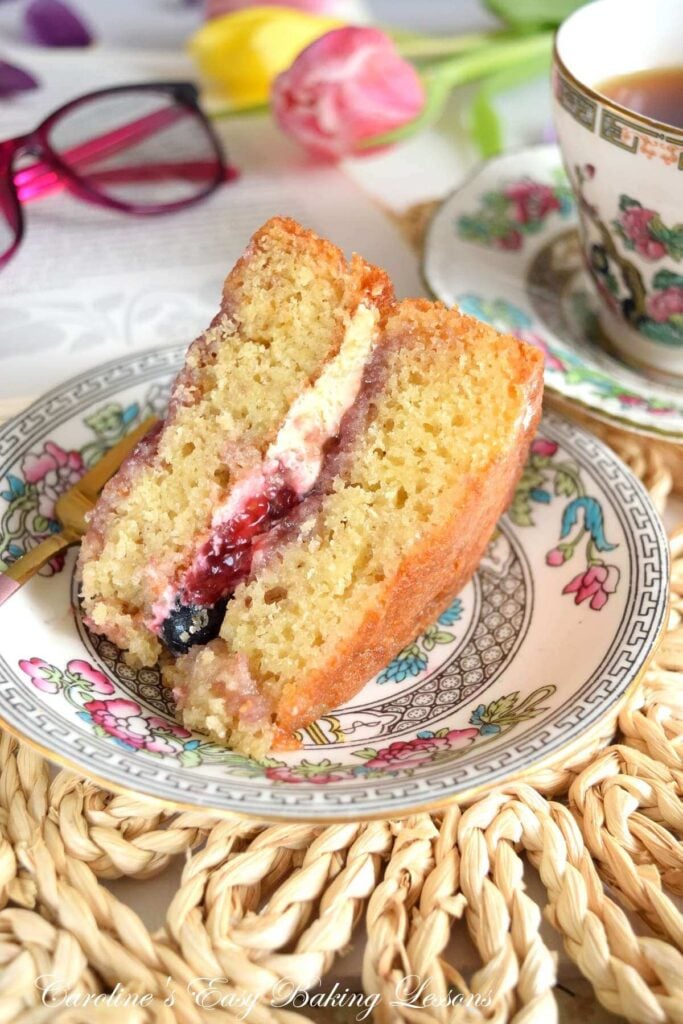Close shot of a slice of raspberry ripple Victoria sandwich cake, next to a book, reading glasses and tea cup.