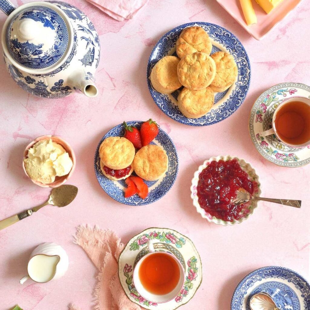Overhead square image of a table with cream tea of vintage bloe willow teapot, cups of tea, napkins, rose, milk, clotted cream, jam & British scones.