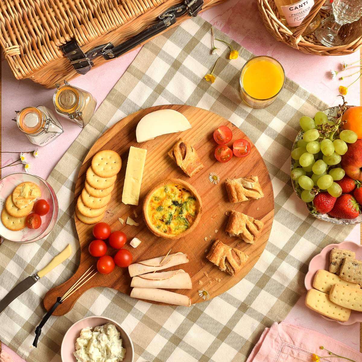 Overhead flatlay image of a picnic setting, with picnic basket, wooden board of sausage rolls, meat slices, tomatoes, cheese, potato salad, biscuits, crackers, fruit & bottle of bubbly.