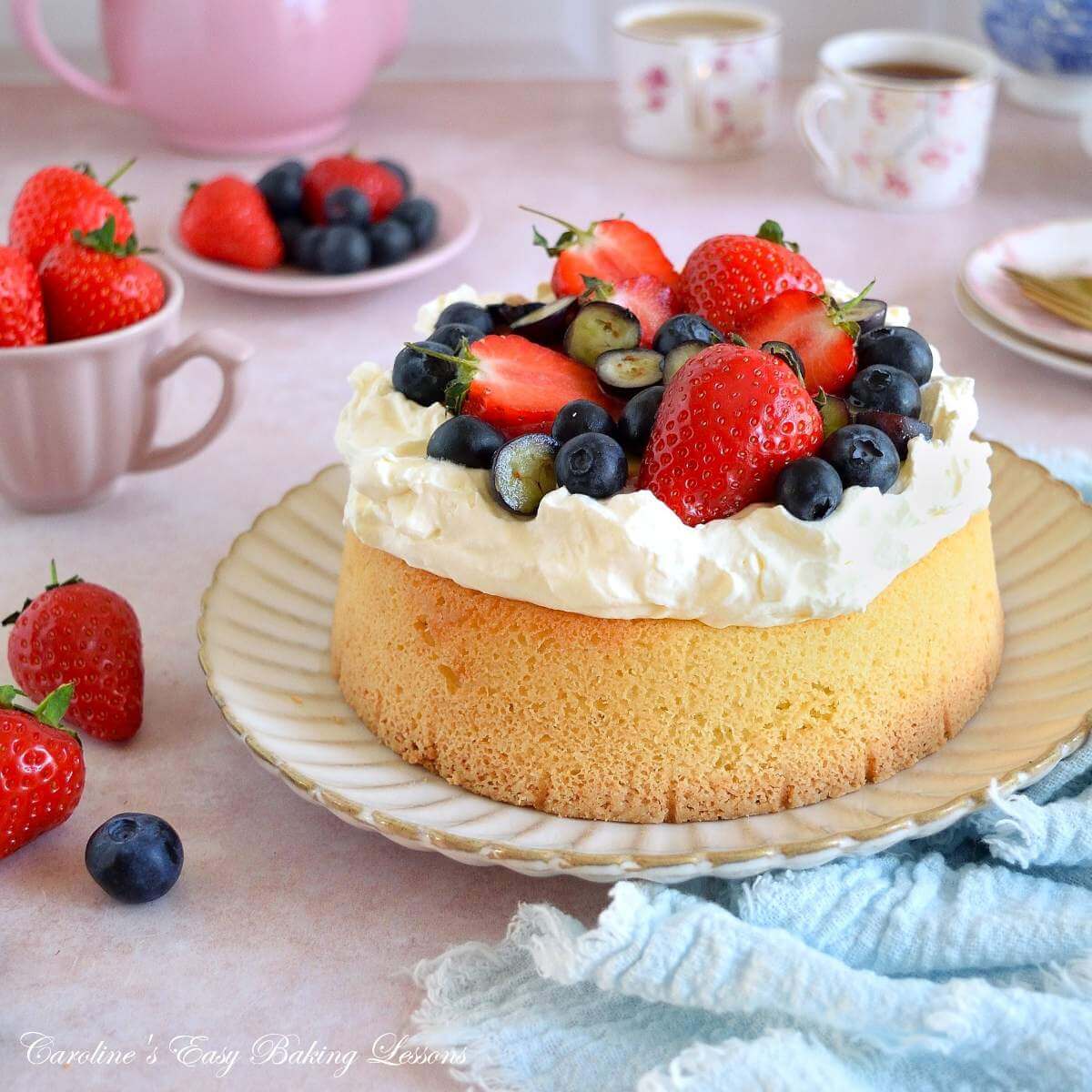 Extra close shot of table with crockery and tea, and a deep cake topped with cream and strawberries and blueberries.