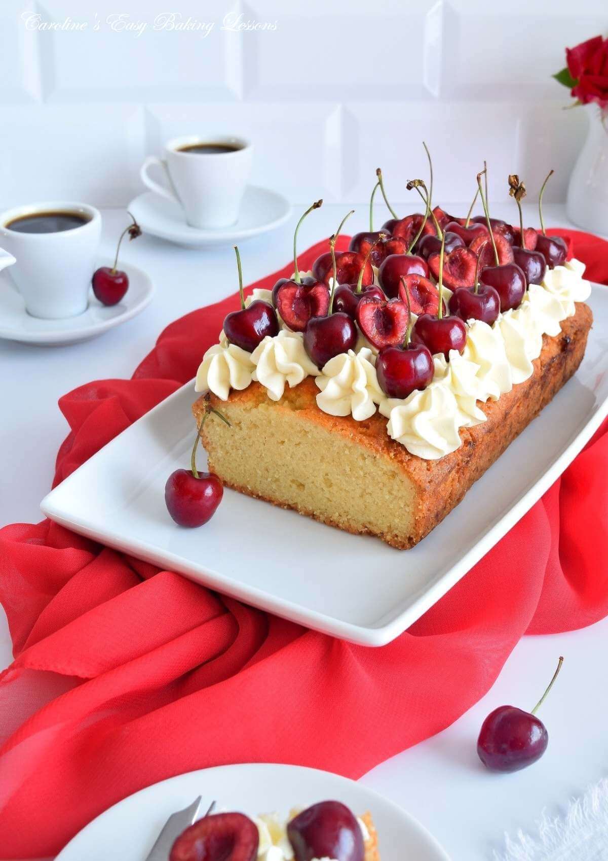 Front on shot of white table with white crockery, coffee cups, backdrop and Madeira loaf cake with piped cream & cherries on top, surrounded by a red large napkin.