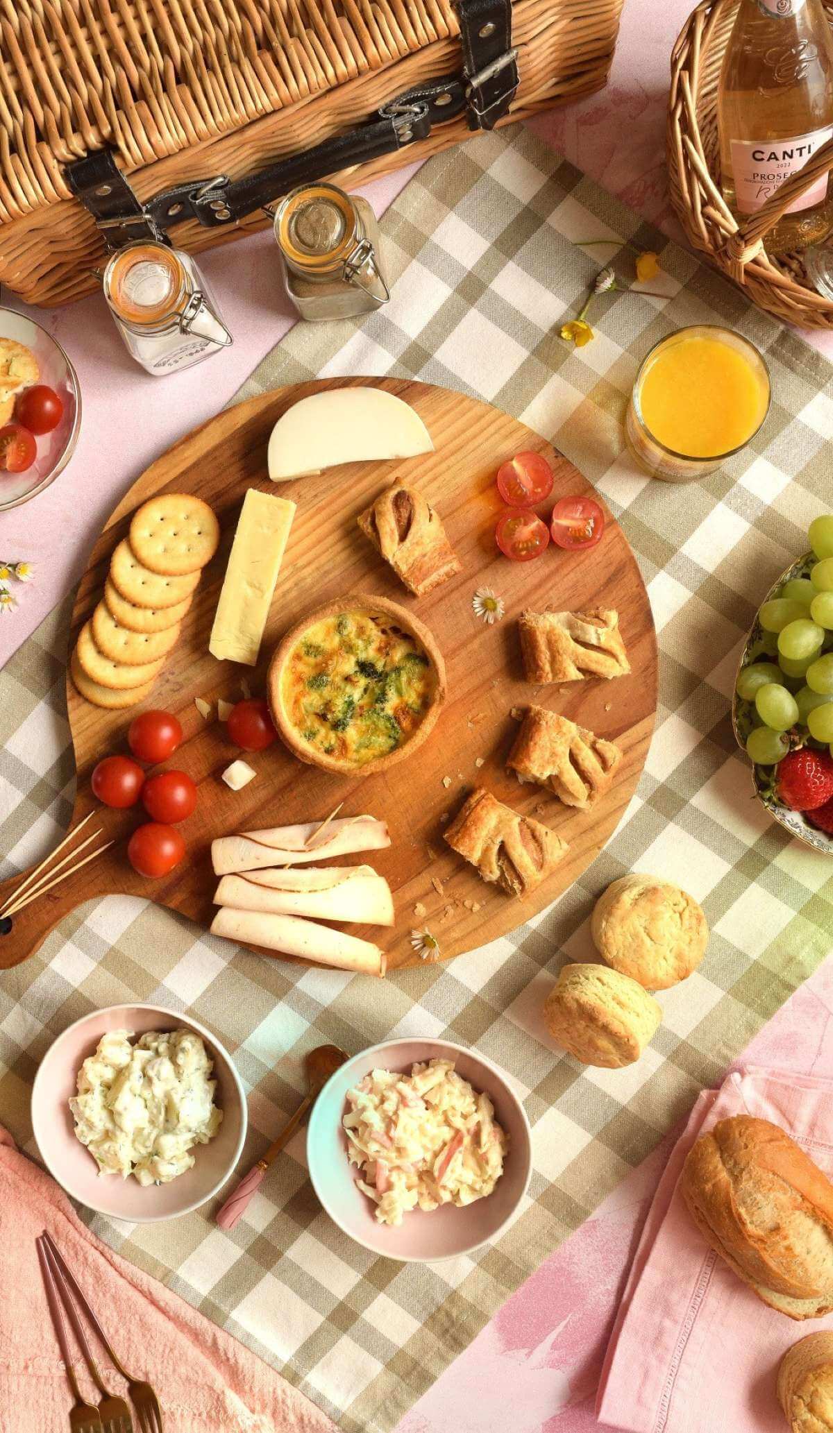 Overhead long flatlya image of a picnic blanket, with picnic basket, basket of bubbly and glasses, juice, fruit, quicke, sausage rolls, crackers, cheese, tomato, potaot salad, coleslay, scones, bread and biscuits.