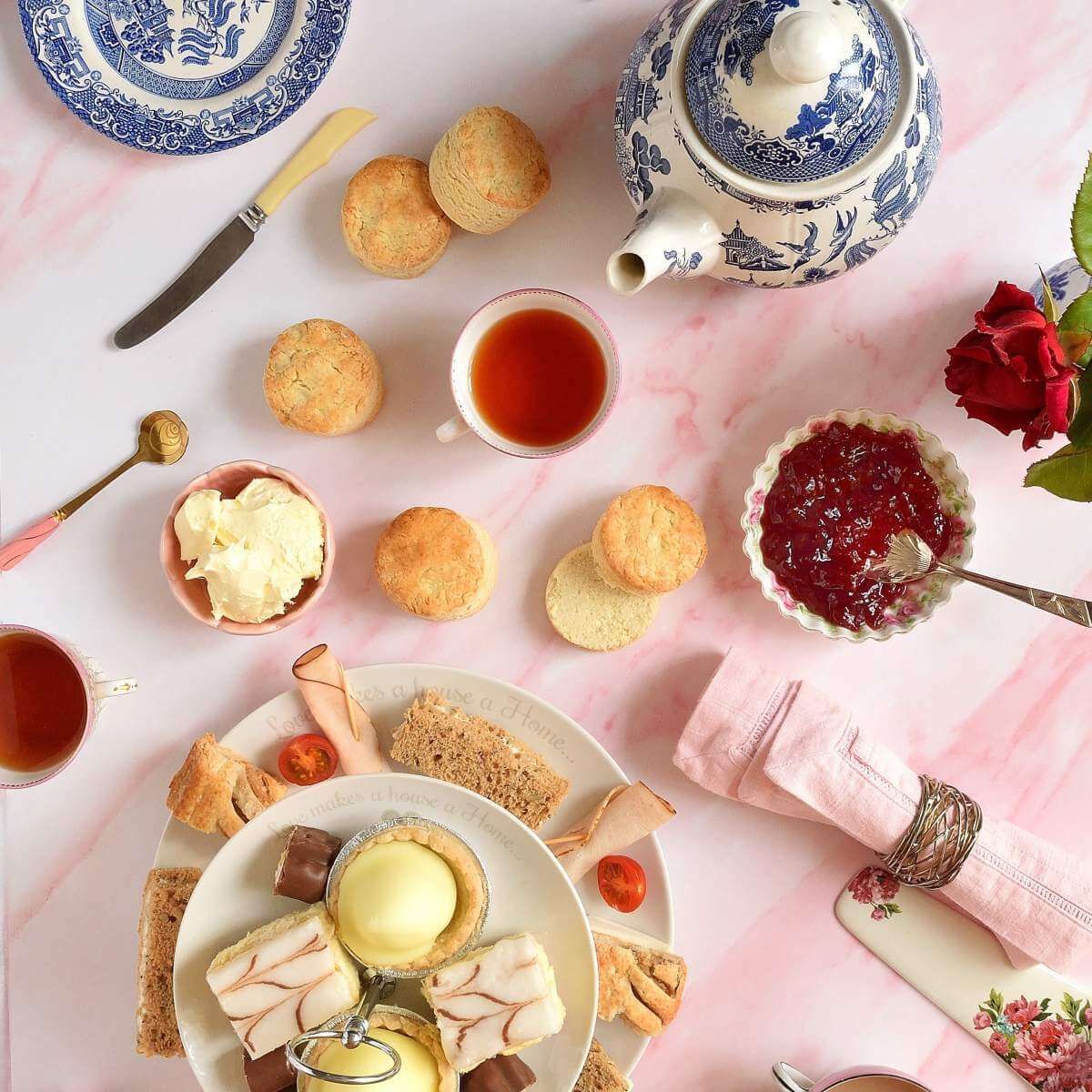 Overhead flat lay image of white pink marble tabletop with afternoon tea setting of scones, tea, cream, jam and cake stand of finger sandwiches and small cakes.
