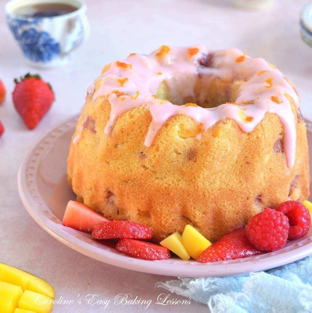 Extra close shot of a golden bundt cake, with pink glaze, mango and strawberry pieces surrounding it.