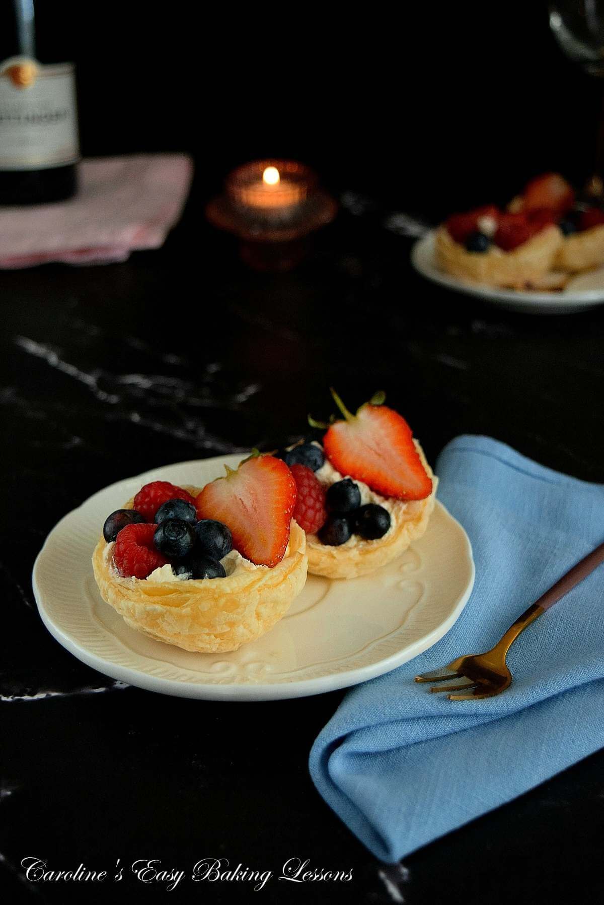 Photo of 2 puff pastry dessert cups, served filled with cream and fresh berries, in a dark restaurant background, with candlelight backing.