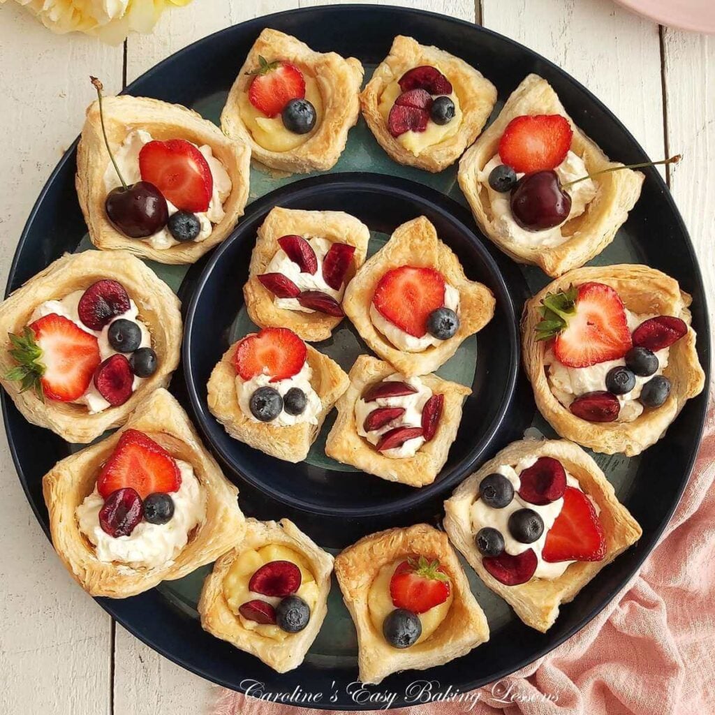 Overhead shot of large round navy blue platter, with 2 circular rows of small an dlarge, puff pastry cups, with cream or lemon curd and fresh berries.