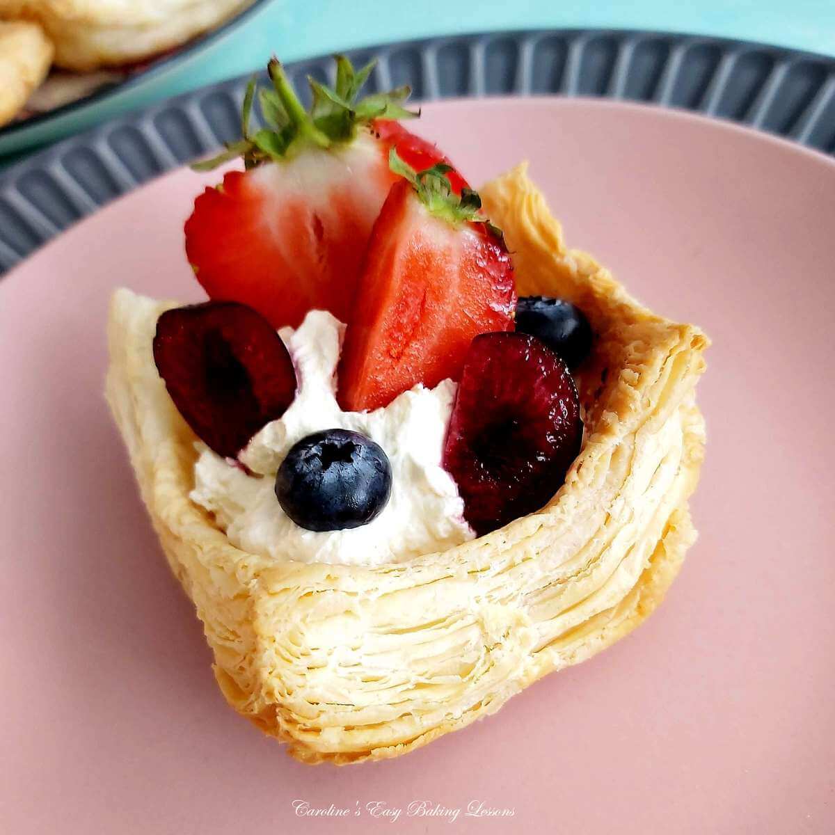 Extra close shot on pink plate, a very flaky puff pastry cup with whipped cream, blueberries, strawberry and halved back cherries.