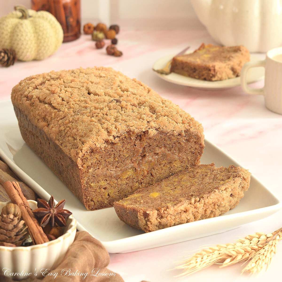 Pink marble counter with platter of pumpkin streusel bread cut, Autumn/Fall decorations, spices & slice served in the background.
