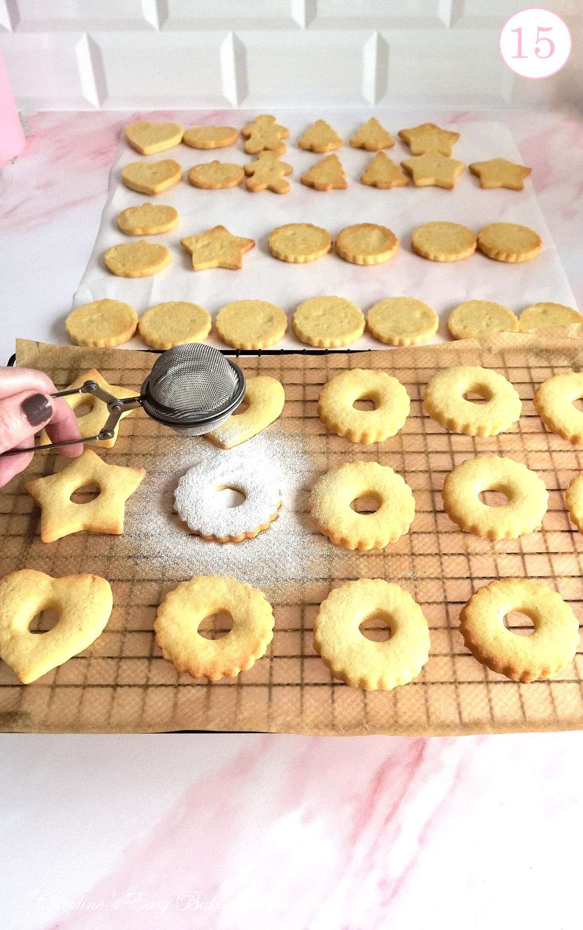 Long shot of seperated shaped cookies, some on cooling rack, with female hand dusting icing/powdered sugar on top, with text 'photo 15'.