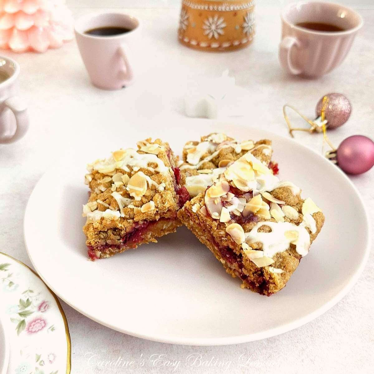 Image on pink table and pink Christmas baubbles and crockery, surrounding a pink plate of 3 staged golden glazed almond topped oat bars with red fresh cranberry filling.