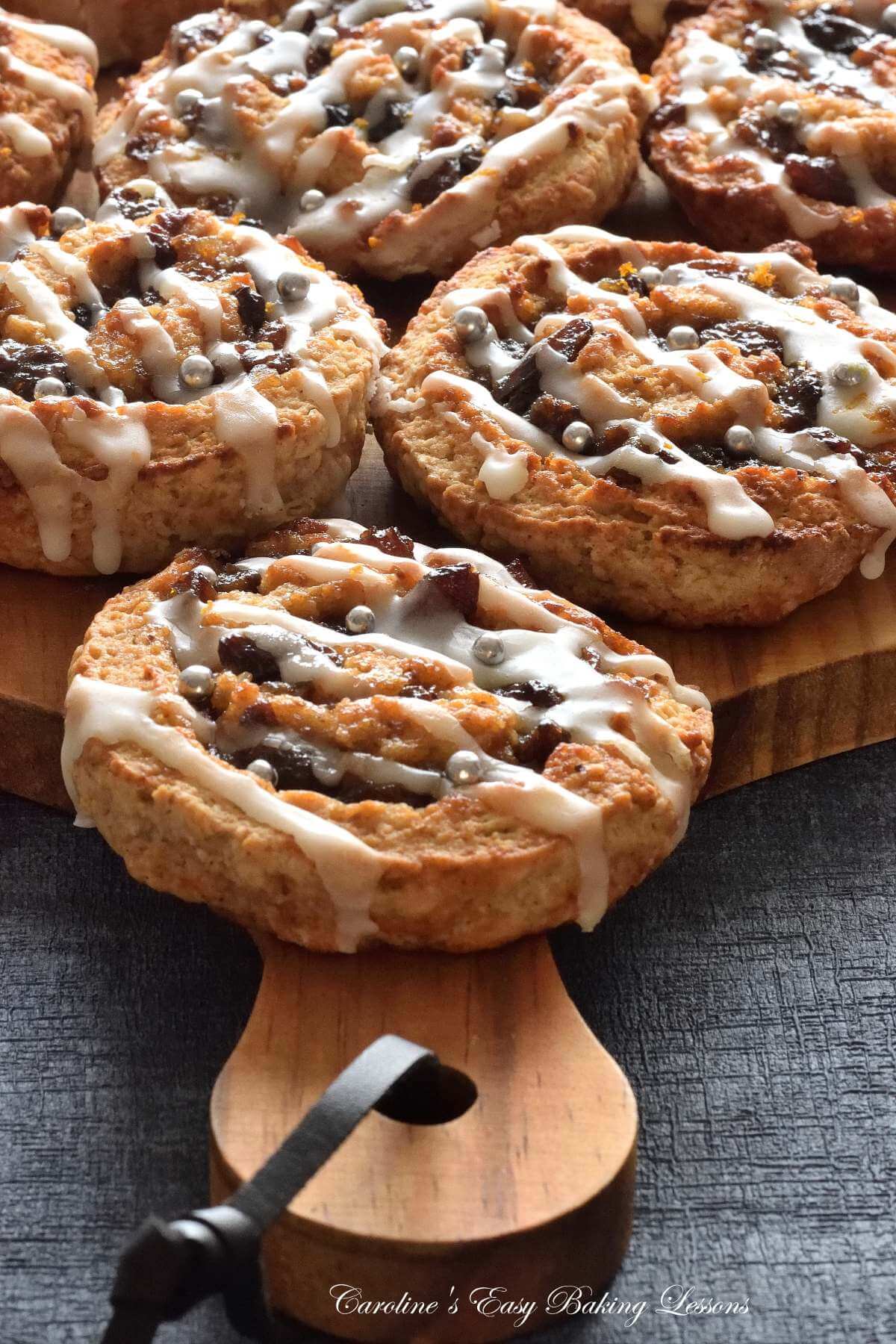 Close, partial shot of several glazed mincemeat swirled buns, on wooden shopping board & navy background.