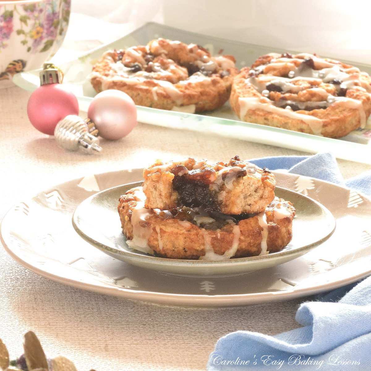 Close shotof Christmas plate with 2 stacked mincemeat filled spiral buns, one one top half eaten, Christmas baubbles to the background.