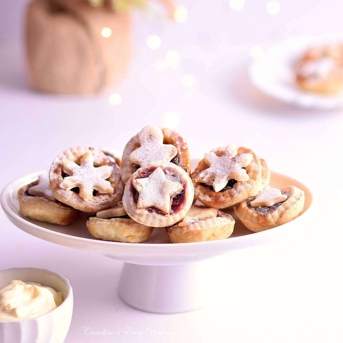 Stack of mini mince pies on white cake stand, white bright setting and fairy lights to the background.