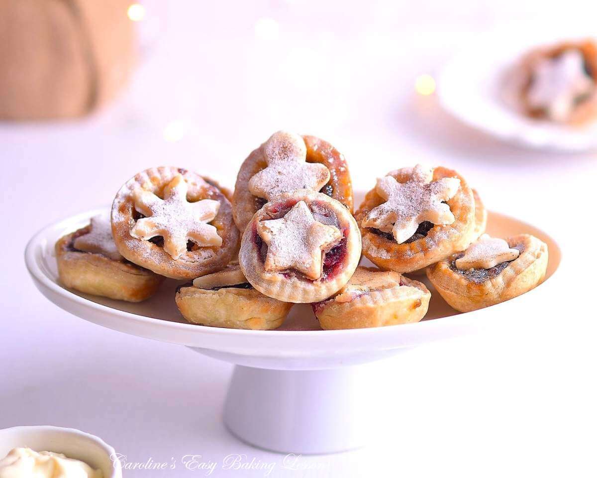 Horizontal, bright airy image of white ovan plate of stacked mini mince pies with seasonal toppers, and white fairy lights in the background.