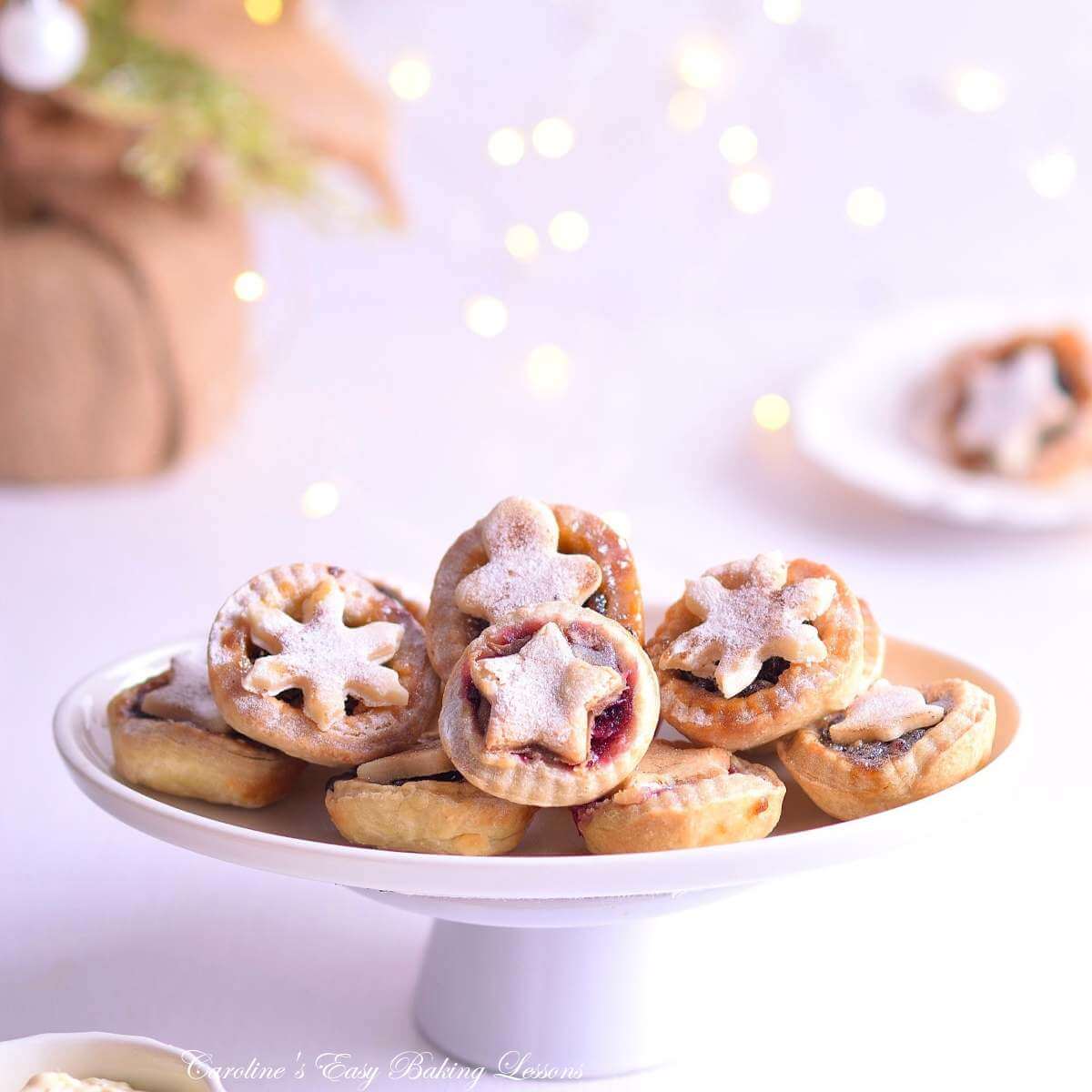 Close shot of a oval white plate on small cake stand, with stack of mini mince pies with seasonal marzipan toppers, white background and fairy lights.