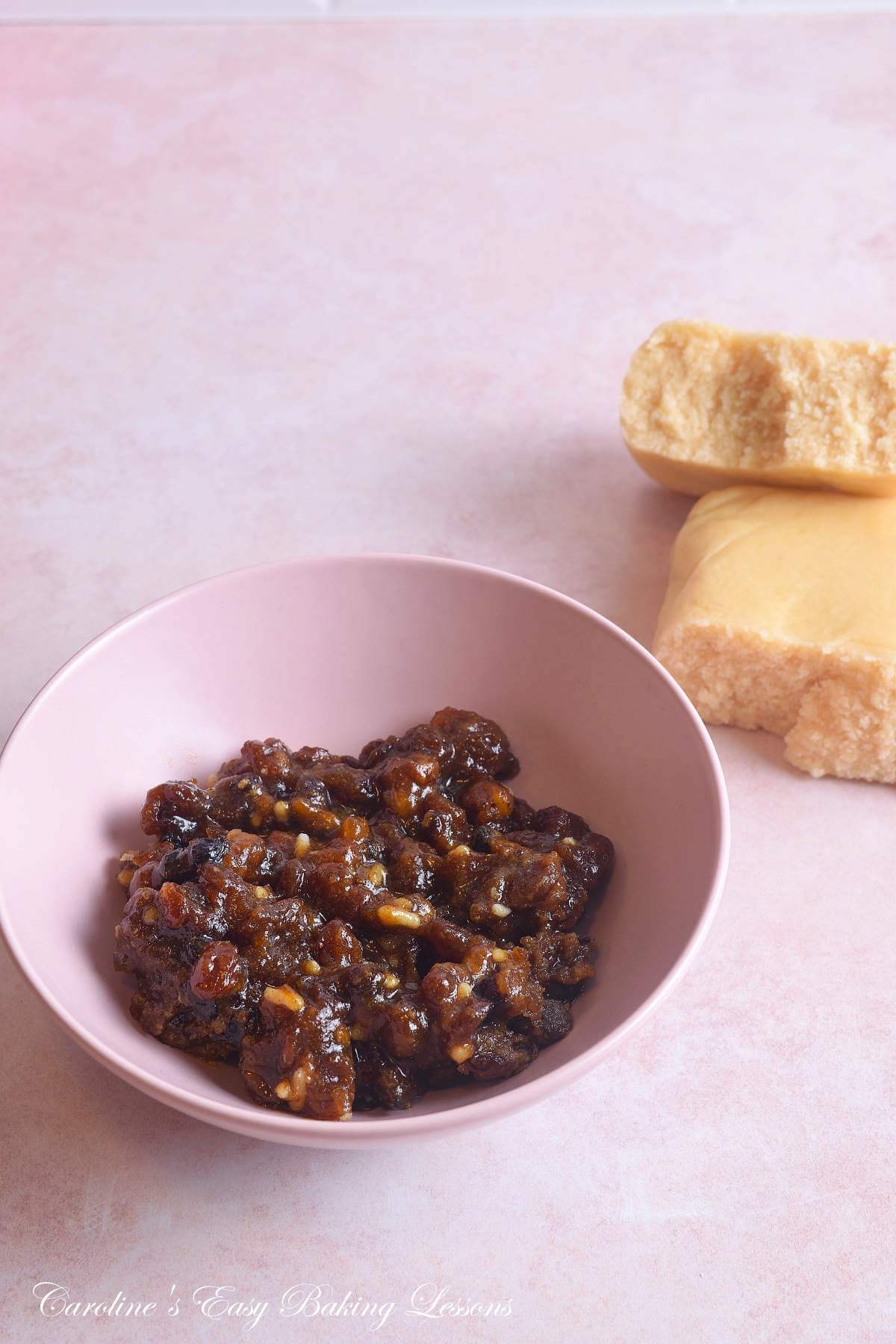 Photo of pink bowl of mincemeat/mince pie filling and halved block of marzipan in the background.