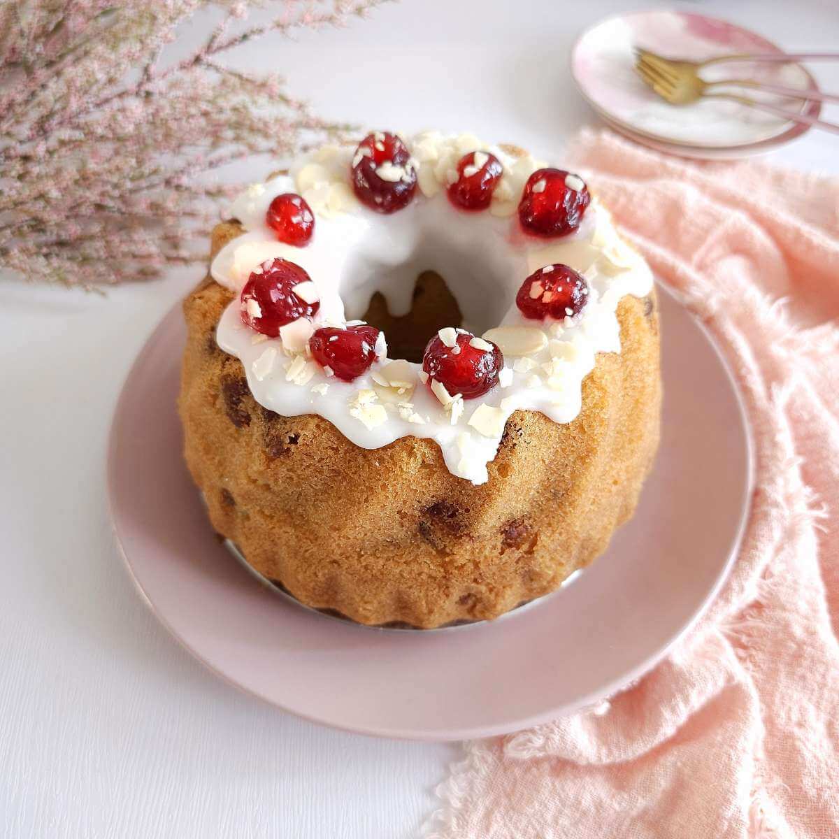 Image of a small Bakewell bundt cake for air fryer, with bright white glaze and cherries, with pink background table.
