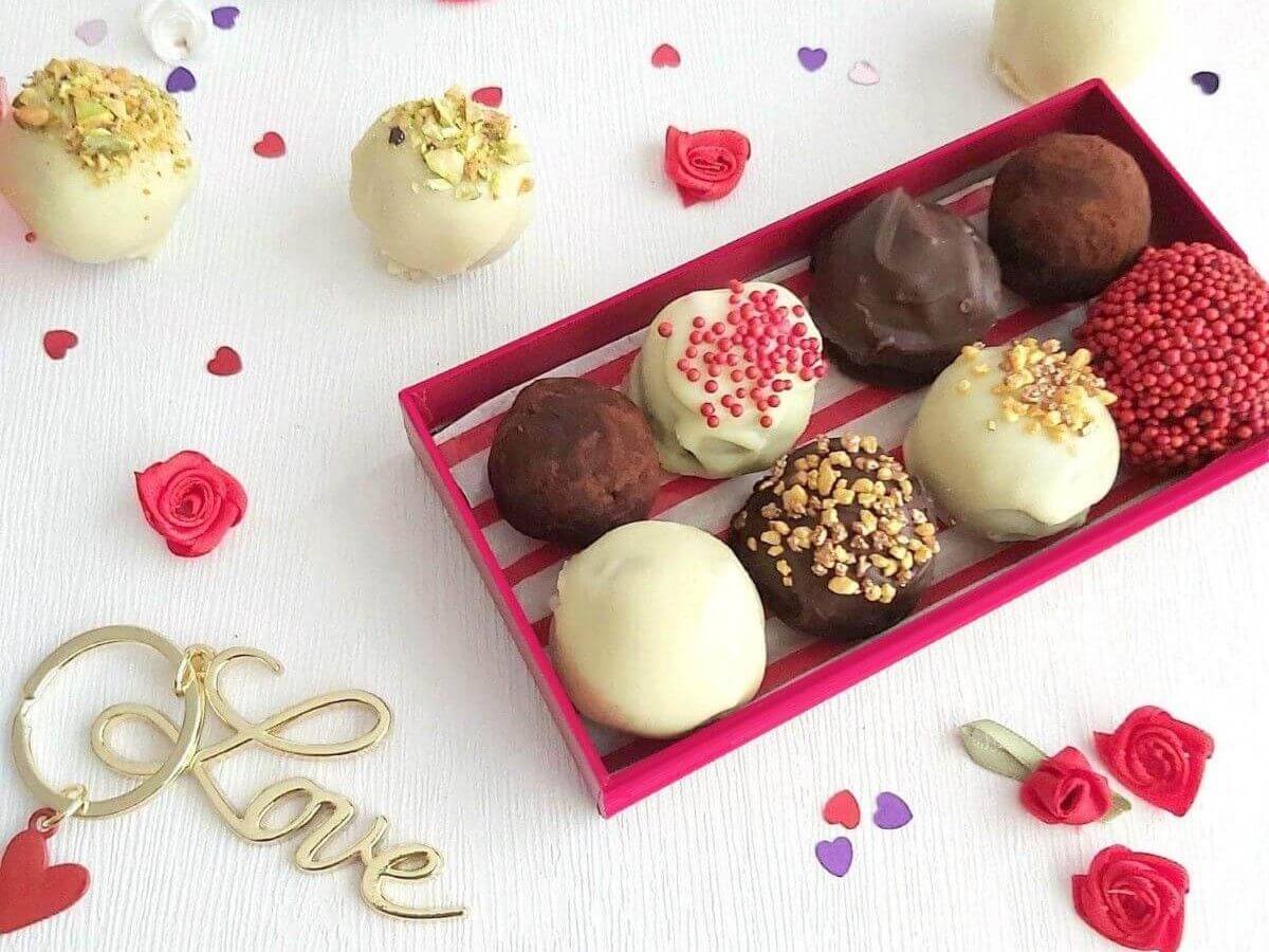 Overhead ofwhite tablewith red box of chocolate coated cake truffles, and hearts and rose confetti surrounding it.