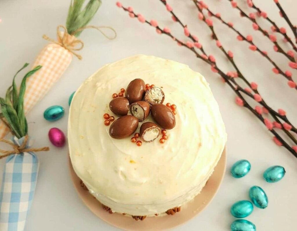 Overhead, horizontal crop image of Easter table with a large carrot cake topped with halves of chocolate Easter eggs.