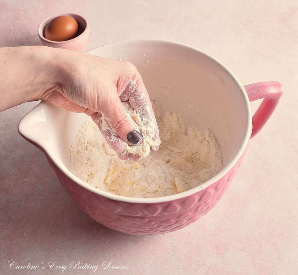 Female hand in pink mixing bowl, 'cutting in' by rubbing butter cubes and flour over fingertips.