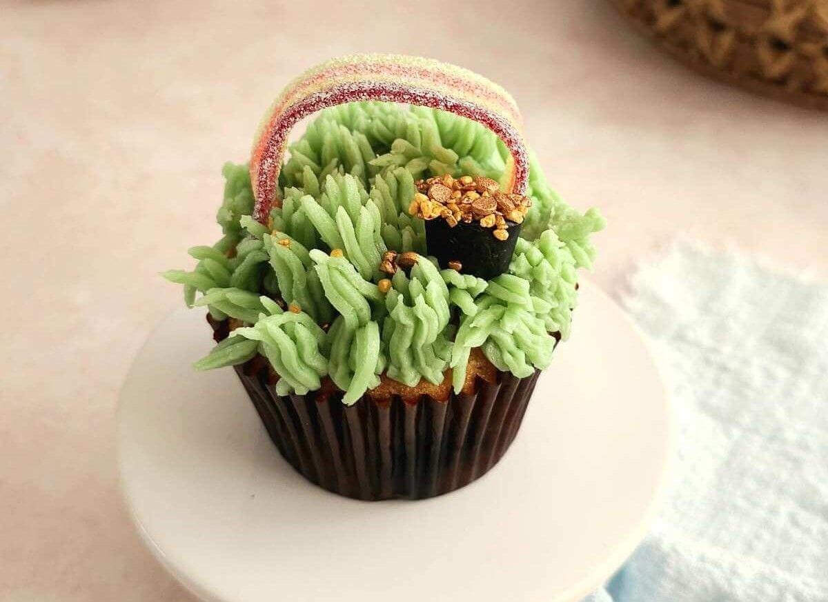 Image of a cupcake with green grass style buttercream, a rainbow candy/sweet on top, and little pots with gold sprinkles.