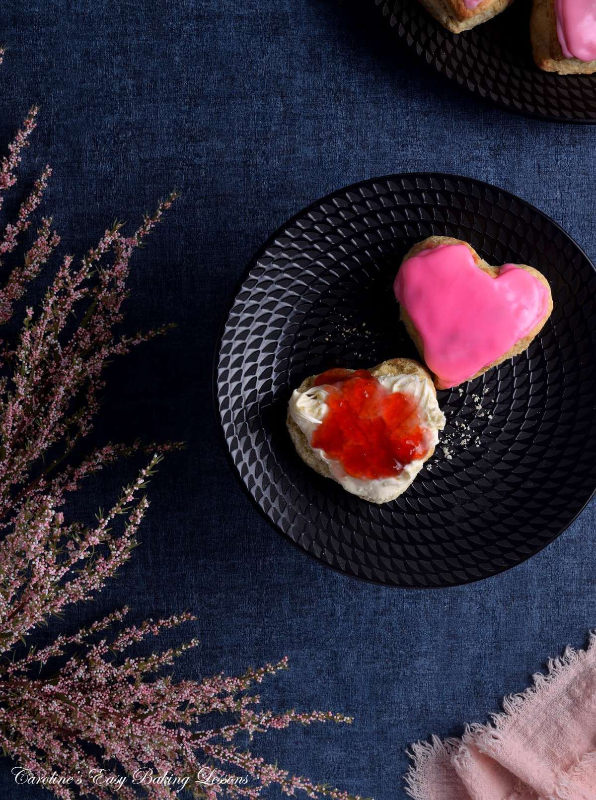 Overhead image, on navy tabletop, with large pink glazed heart-shaped British scone, on black plate, with crema and jam, and pink napkin and folliage framing it.