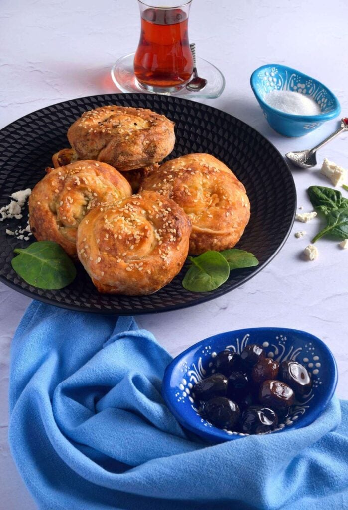 Image of Turkish rolled/rose shaped filo pastries on black plate, with Turkish tea glass, spinach leaves and white cheese surrounding the plate.