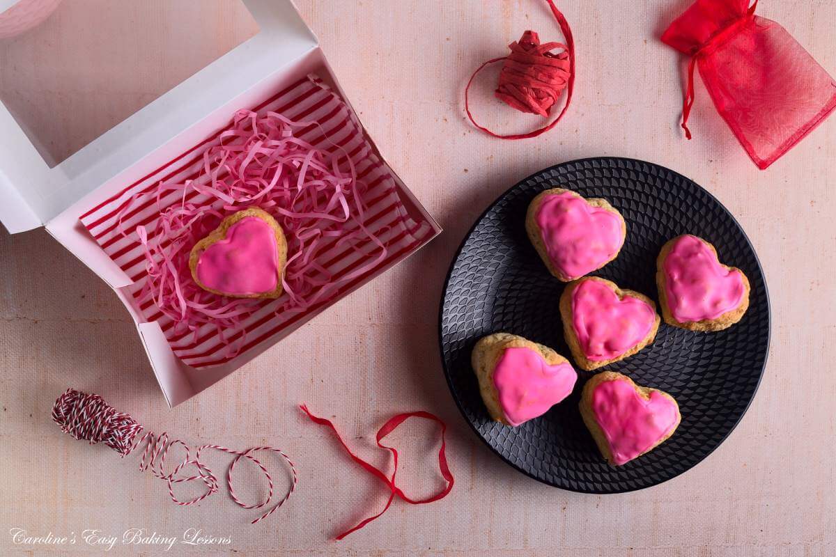 Overhead image on linent ableof black playe holding pink glazed large heart-shaped British scones, one in a cake box, pink shredded paper and red ribbon for gifting.