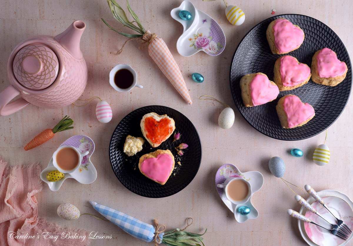 Overhea dimage of tabletop, with pink and lback crockery, with large pink glazed heart-shaped British scones, with Easter decorations and eggs.