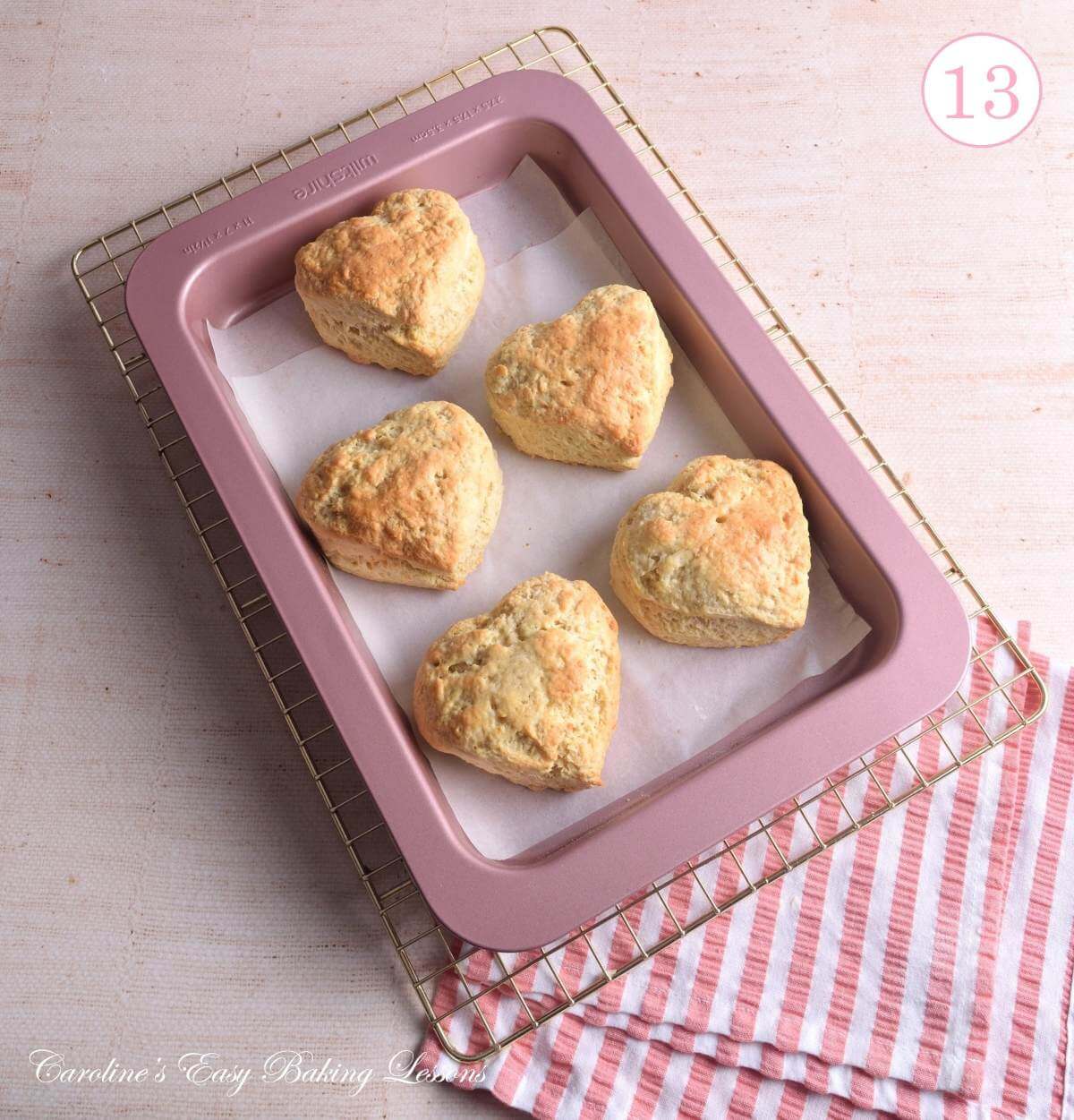 Overhead image on tabletop, of pink/rose gold baking tray, with 5 baked heart-shaped large British scones, on cooling rack, with text 'photo 13'.