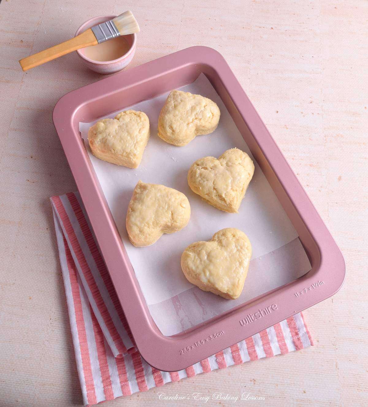 Soft toned 45 degreee angled shot of rose gold baking tray holding 5 large heart-shaped British scones before baking and egg wahs and brush to the side.