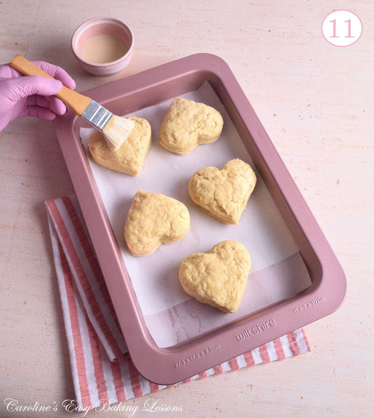 Image of rose gold/pink baking tray with 5 large heart-shaped British scones befor ebaking, pink gloved hand brushing on egg-wash and text 'photo 11'.