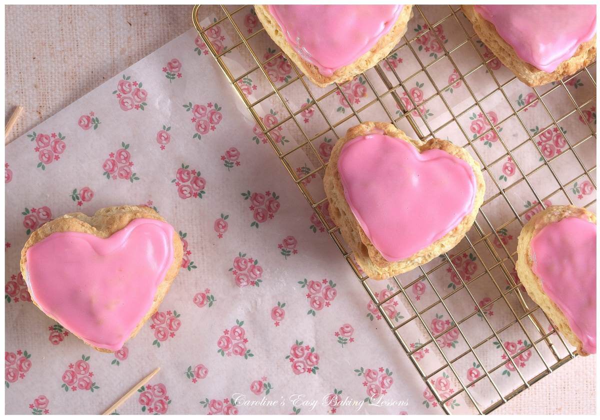 Overhead, partial image, clos eup of pink rose bakign paper, large pink glazed heart shaped British scones, showing glaze setting.