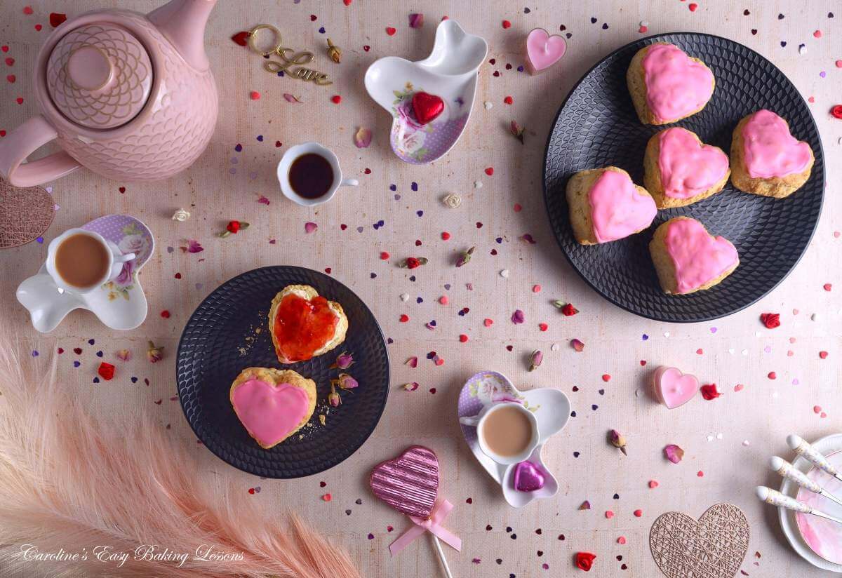 Overhead flat lay image on beigh table, with pink heart shaped British scones, one with crema and jam, crockery and lot sof heart sprinkles and heart chocolates for Valentine's.