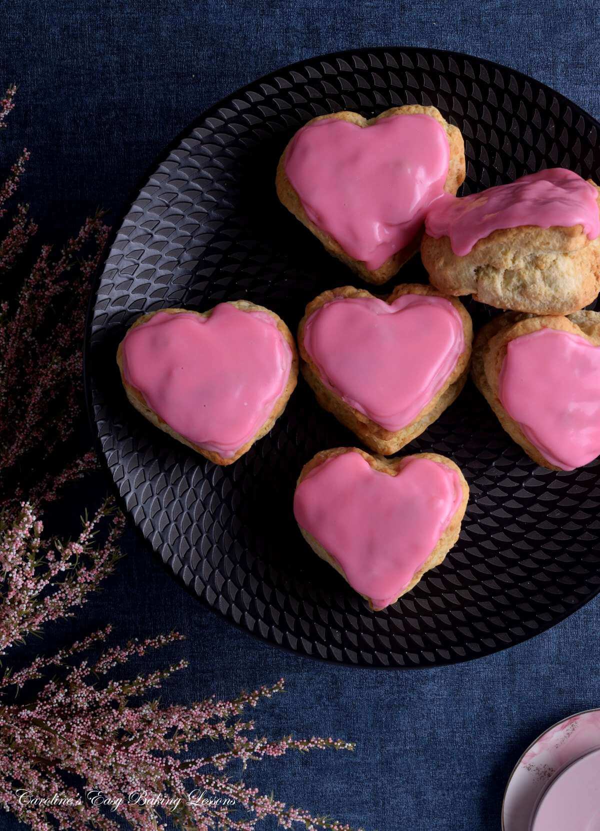Close overhead image of navy tabletop, with black plate partially showing pink glazed large heart-shaped British scones.
