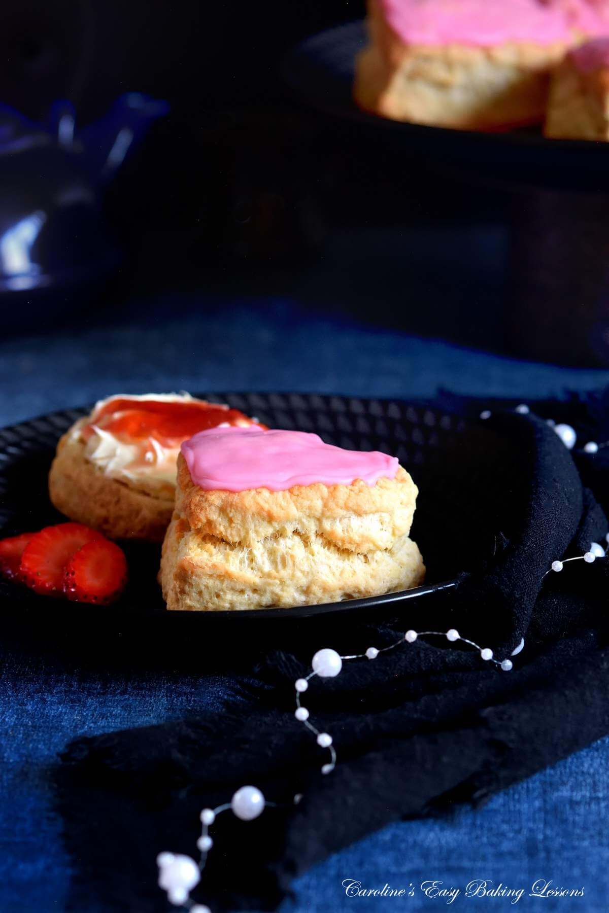 Dark image on navy backdrop of black plate with pink heart shaped British scone, one with cream and jam and more on a cake stand in the background.