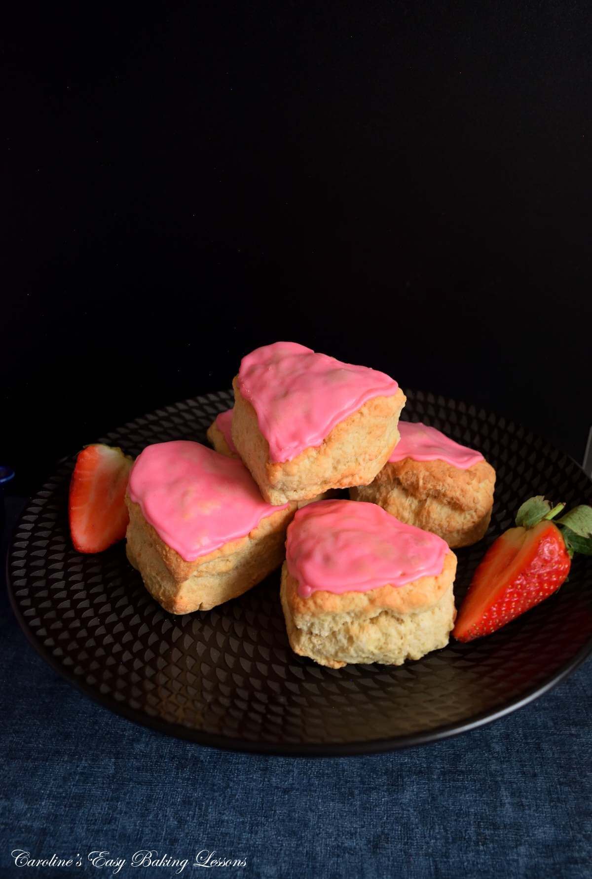 Front angled image on navy tabletop and black background, black cake stand with stack of large, pink glazed, heart-shaped British scones, and strawberry halves.