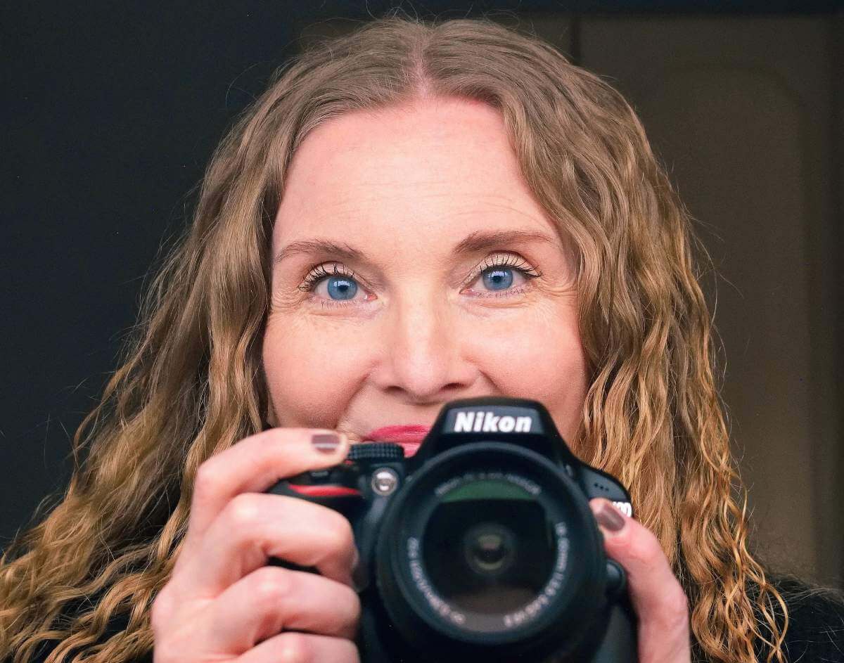 Close front on image of red curly haired 50 something female holding up Nikon dslr camera.