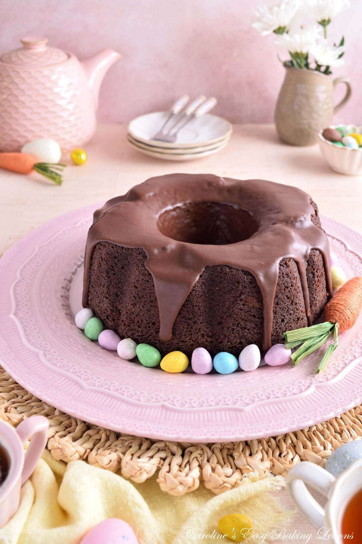 Vertical image of pastel/Spring coloured table with crockery, linen and Easter decorations, around a Devil's Food chocolate bundt cake with mini eggs.