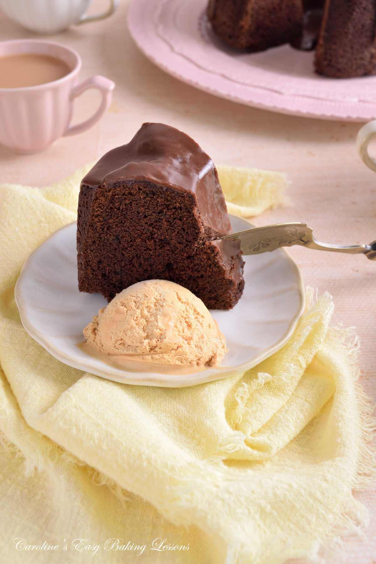 Vertical image of a pastel table with a slice of Devil's Food chocolate bundt cake served with icecream and a spoon starting to eat.