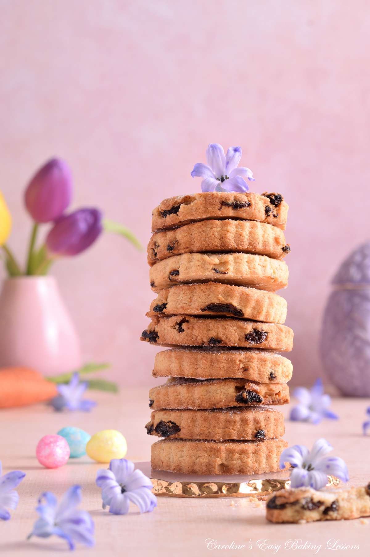 Close shot of a 9 cookie stack of Mary Berry Easter biscuits, with hyacinth flower on top, at the base and pastel mini eggs surrounding.