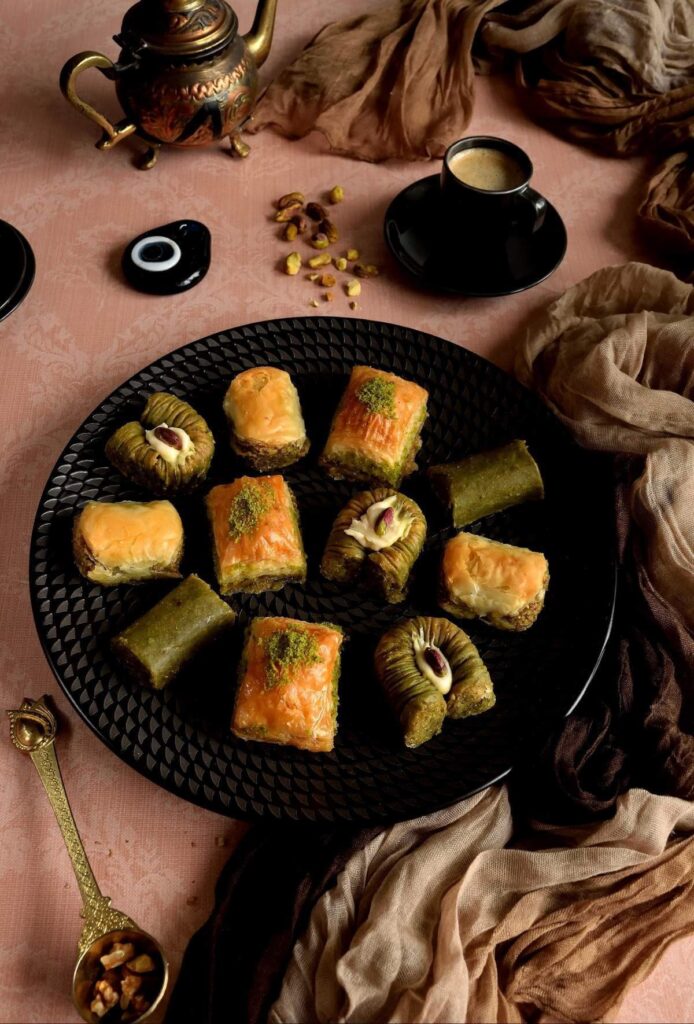 45 degree angled dark shot of wamr coloured table top with brown fabric, Turkish coffee, props surrounding a black plate of fancy Turkish Baklava by Hacci Hakki Usa brand.