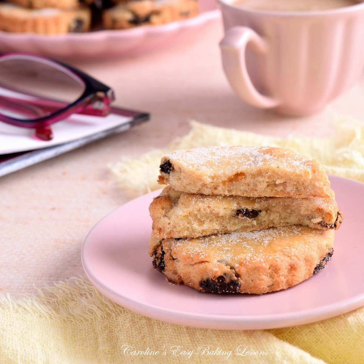 Close shot of a scene of pastel coloured tea, crockery, corner of a book and reading glasses and a stack of 2 Easter biscuits/cookies.
