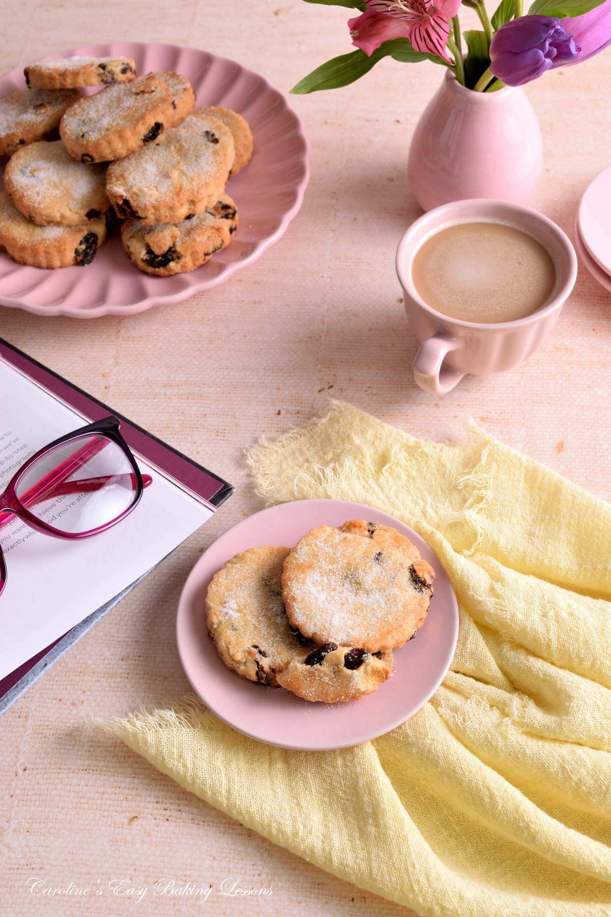 45 degree angeled shot of apstel coloured table with linene and crockery, flower, coffee, book and reading glasses surrounding a plate of Easter biscuits/cookies.