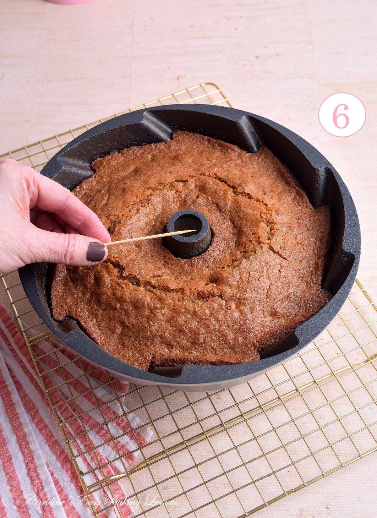 Image of female hand holding clean cocktail stick in front of a baked gluten free carrot bundt cake cooling, with text 'photo 6'.