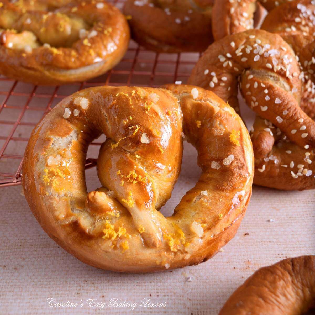 Extra close shot of a sweet, orange syrup coated & zest, homemade yeast pretzel, with more in the background.