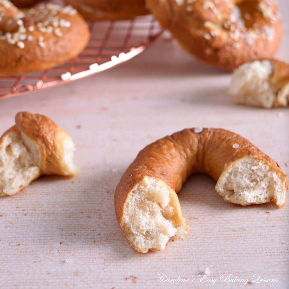 Clos eimage of a piece of pretzel, showing the soft white fluffy texture inside chewy brown crust, with some whole pretzels to the far background.