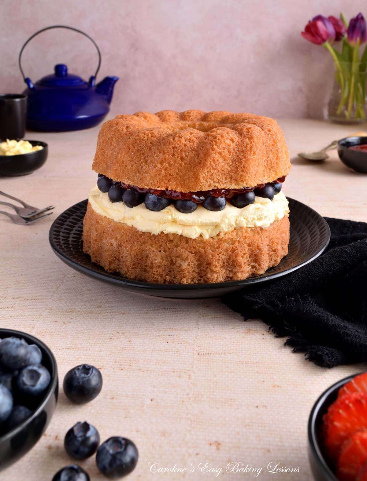 Image of a golden Victorian Sandwich layered bundt cake on black plate, with crockery and berries to the side, but filling of cream, bluberries and jam being prominant.